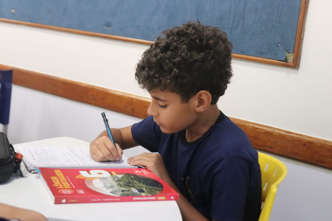 Aluno fazendo a tarefa no caderno com o livro de Língua Portuguesa aberto