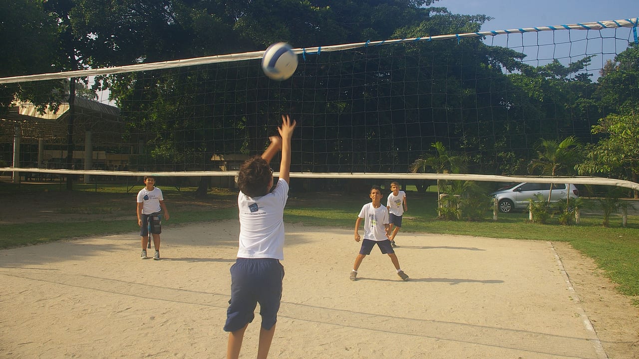 Aluno tocando a bola por cima da rede em aula de vôlei na quadra de areia